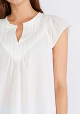 [Color: White] A close up front facing image of a brunette model wearing a white bohemian top crafted from lightweight cotton gauze. With short cap sleeves, a split v neckline, gathered details at the yoke, and a raw edge hem. 
