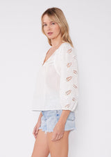 [Color: White] A side facing image of a blonde model wearing a white cotton bohemian blouse. With three quarter length sleeves, elastic wrist cuffs, a split v neckline with ties, embroidered sleeves, lace trim, and pintuck details.