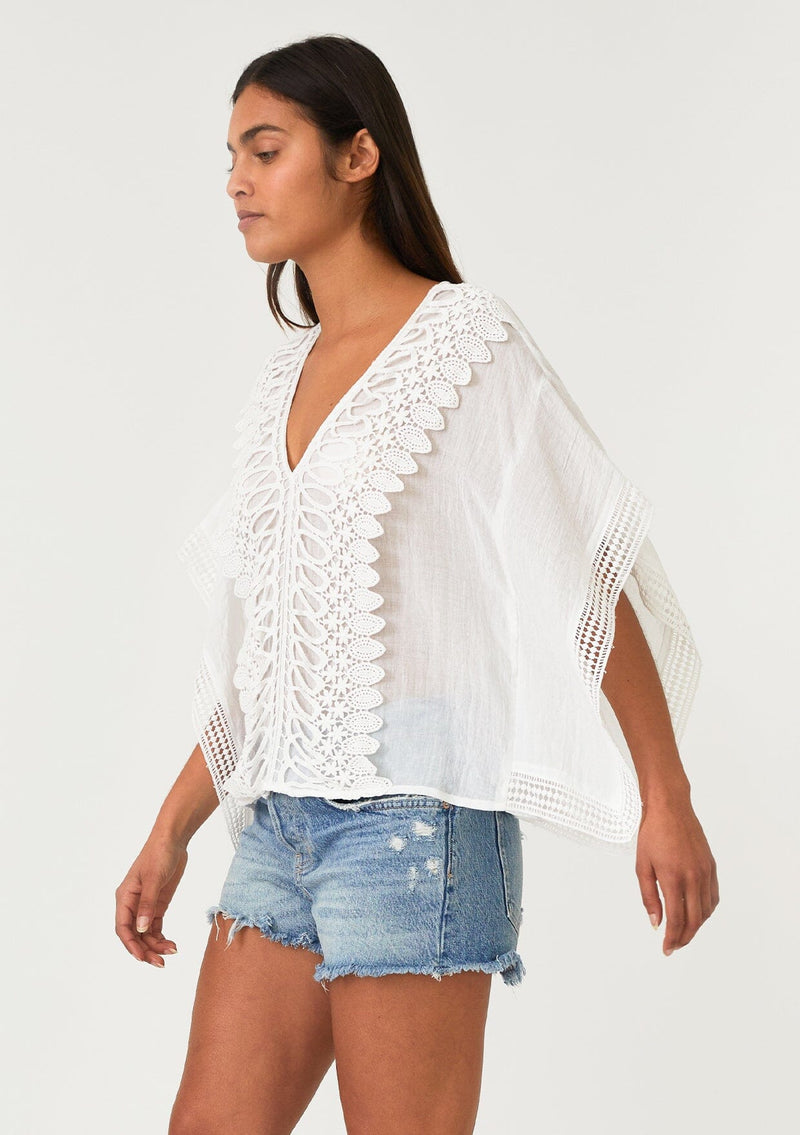 [Color: Off White] A side facing image of a brunette model wearing a sheer white bohemian beach cover up top. With crochet lace trim, short sleeves, a v neckline, and a relaxed, boxy fit. Perfect for the beach or wearing over your swimsuit on your next vacation. 