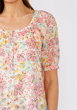 [Color: Natural/Peach Blossom] A close up front facing image of a brunette model wearing a pink floral cotton summer blouse. With short puff sleeves, a scooped neckline, and a button front. 