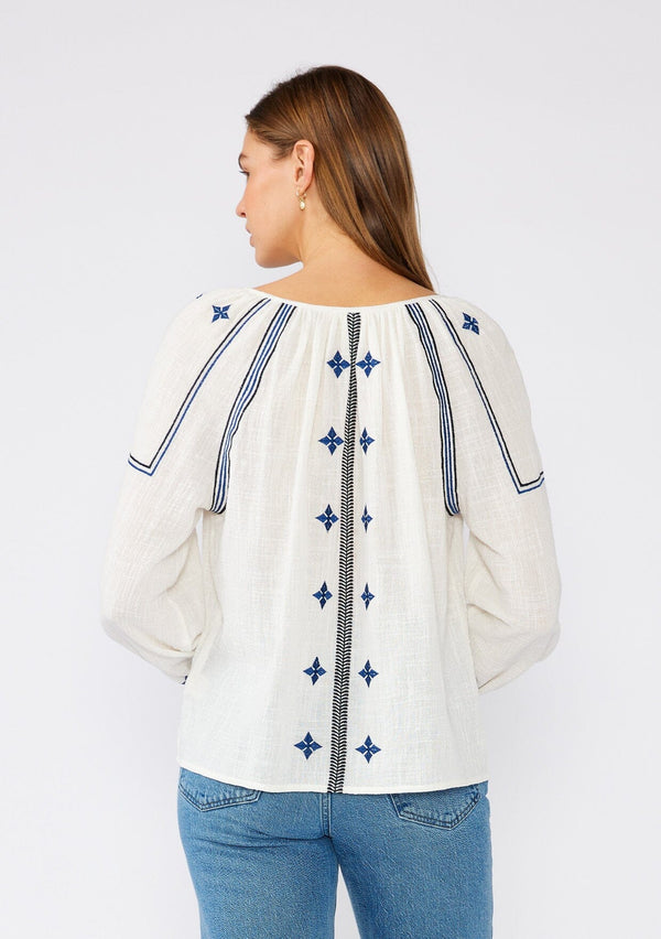 [Color: White/Navy] A back facing image of a brunette model wearing a white cotton bohemian blouse with navy blue embroidered details. With long sleeves, a round neckline, a button front, and a relaxed fit. 