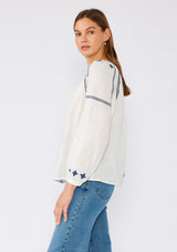 [Color: White/Navy] A side facing image of a brunette model wearing a white cotton bohemian blouse with navy blue embroidered details. With long sleeves, a round neckline, a button front, and a relaxed fit. 