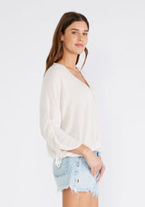 [Color: Almond] A side facing image of a brunette model wearing a light ivory bohemian blouse with three quarter length sleeves, a drawstring sleeve with tie accent, a v neckline, a self covered button front, and crochet lace trim. 