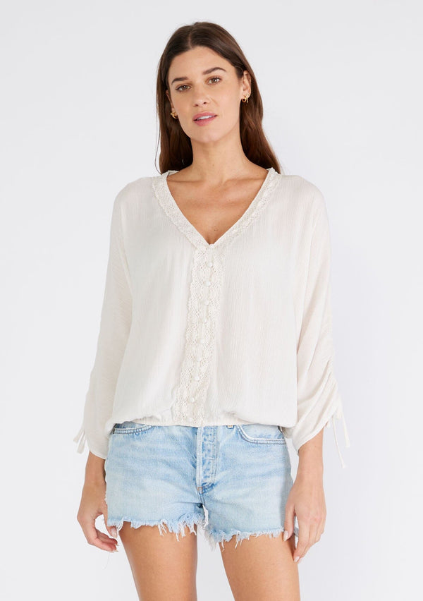 [Color: Almond] A front facing image of a brunette model wearing a light ivory bohemian blouse with three quarter length sleeves, a drawstring sleeve with tie accent, a v neckline, a self covered button front, and crochet lace trim. 