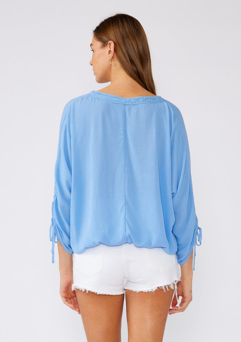 [Color: Periwinkle] A back facing image of a brunette model wearing a blue bohemian blouse with three quarter length sleeves, a drawstring sleeve with tie accent, a v neckline, a self covered button front, and crochet lace trim. 