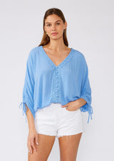 [Color: Periwinkle] A front facing image of a brunette model wearing a blue bohemian blouse with three quarter length sleeves, a drawstring sleeve with tie accent, a v neckline, a self covered button front, and crochet lace trim. 