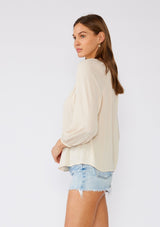 [Color: Natural/Pink] A side facing image of a brunette model wearing a cream colored bohemian blouse with floral embroidered details. With long raglan sleeves, a v neckline, a button front, and contrast stitch details along the hemline. 