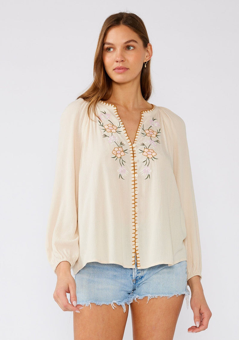 [Color: Natural/Pink] A front facing image of a brunette model wearing a cream colored bohemian blouse with floral embroidered details. With long raglan sleeves, a v neckline, a button front, and contrast stitch details along the hemline. 