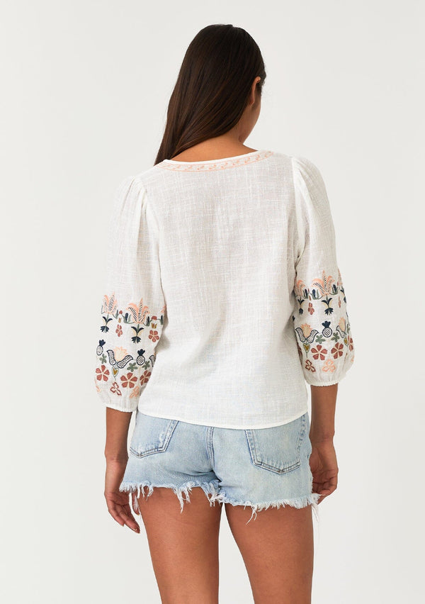 [Color: Natural/Coral] A back facing image of a brunette model wearing an off white bohemian blouse with colorful floral embroidered details throughout. Featuring three quarter length sleeves, a v neckline, and a tie waist detail. 