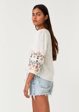[Color: Natural/Coral] A side facing image of a brunette model wearing an off white bohemian blouse with colorful floral embroidered details throughout. Featuring three quarter length sleeves, a v neckline, and a tie waist detail. 