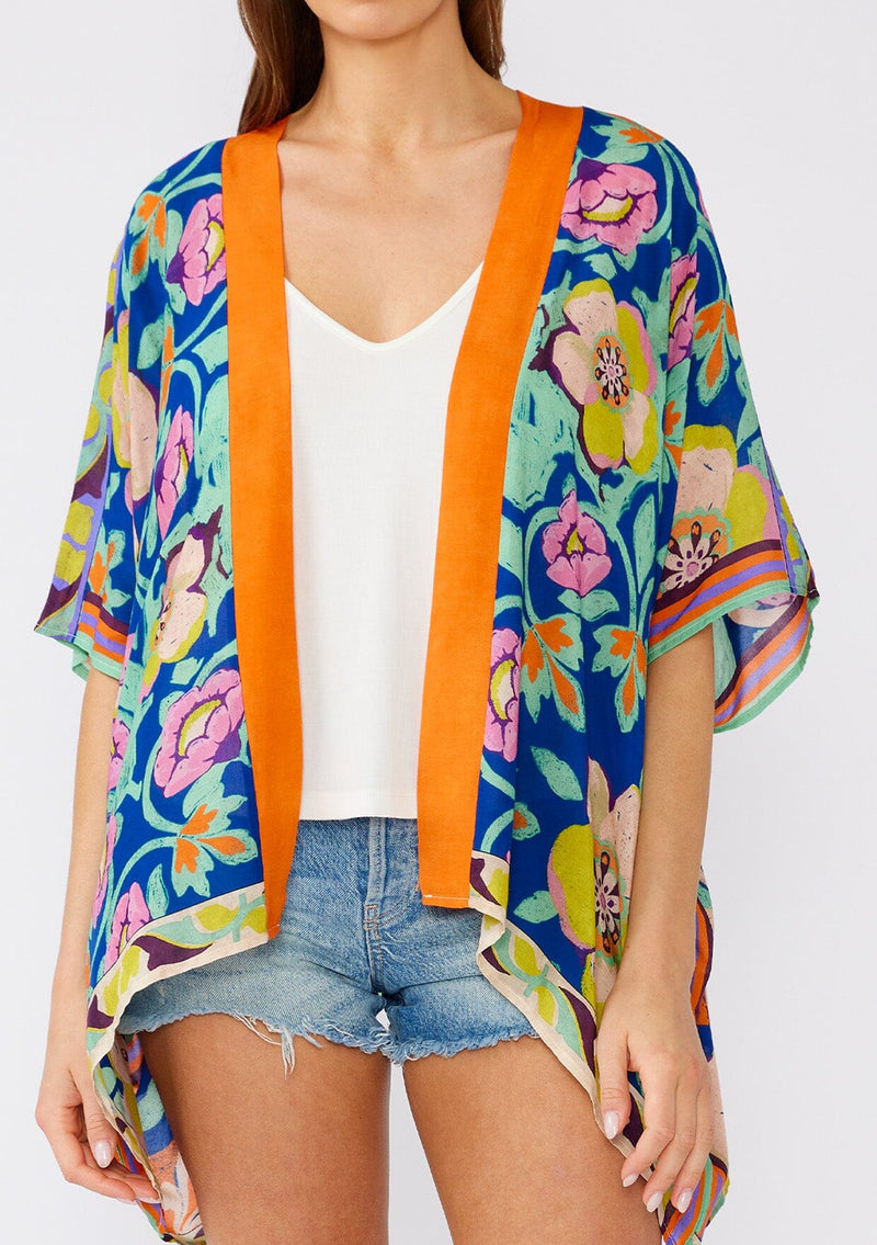 [Color: Blue/Coral] A close up front facing image of a brunette model wearing a bohemian style kimono top in a bright blue and coral floral print with a contrast orange border. A lightweight beach cover up with half length kimono sleeves, an open front, and a hip length hemline. 