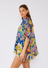 [Color: Blue/Coral] A side facing image of a brunette model wearing a bohemian style kimono top in a bright blue and coral floral print with a contrast orange border. A lightweight beach cover up with half length kimono sleeves, an open front, and a hip length hemline. 