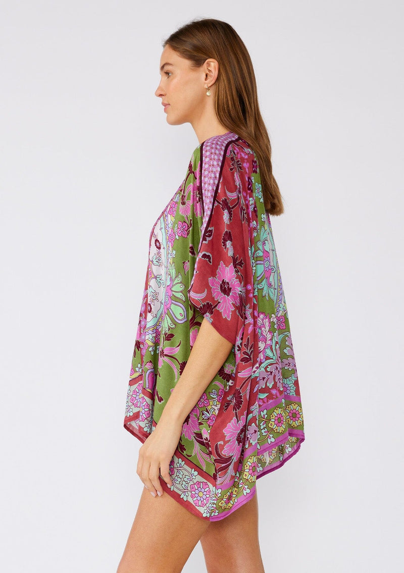 [Color: Green/Rose] A side facing image of a brunette model wearing a bohemian style kimono top in a green and pink floral print, with contrast border. A lightweight beach cover up with half length kimono sleeves, an open front, and a hip length hemline. 