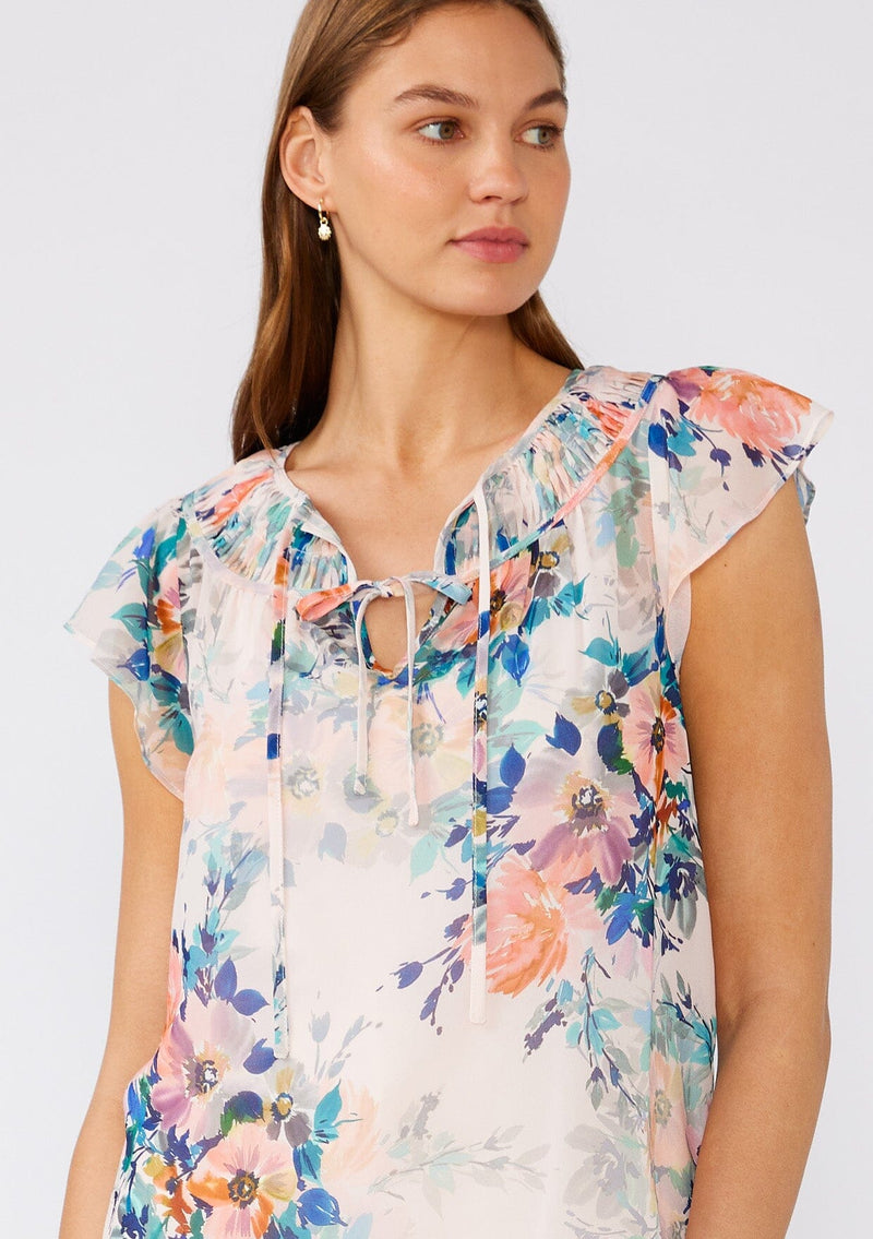 [Color: Peach/Teal] A close up front facing image of a brunette model wearing a sheer chiffon top in a pink and blue floral print. With short flutter sleeves, a split v neckline with double ties, and pintuck details along the neckline. 