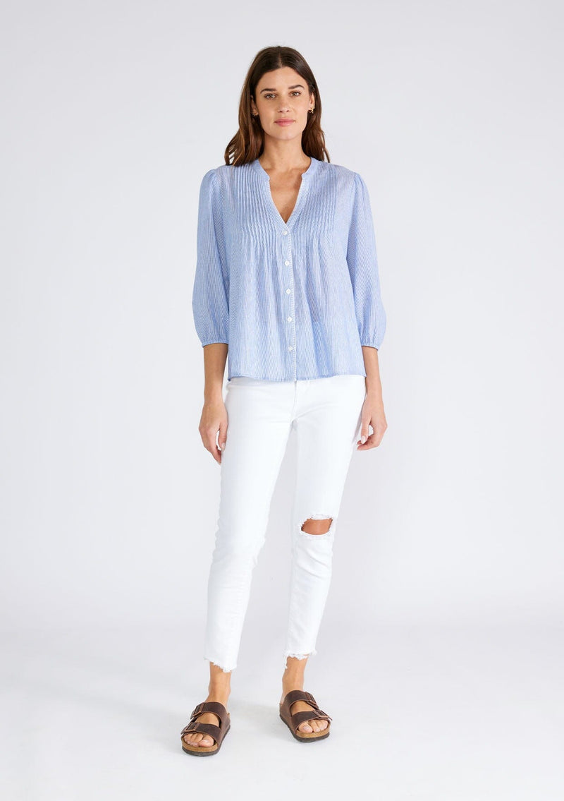 [Color: Chambray/Ivory] A full body front facing image of a brunette model wearing a classic cotton blouse in a blue and white pinstripe. With three quarter length sleeves, a split v neckline, a button front, pleated pintuck details, and contrasting blanket stitch details. The flowy fit is both comfortable and chic for spring. 