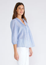 [Color: Chambray/Ivory] A side facing image of a brunette model wearing a classic cotton blouse in a blue and white pinstripe. With three quarter length sleeves, a split v neckline, a button front, pleated pintuck details, and contrasting blanket stitch details. The flowy fit is both comfortable and chic for spring. 