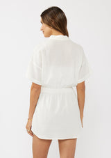 [Color: White] A back facing image of a brunette model wearing a white tunic shirt with a collared neckline, a button front, short sleeves with a dropped shoulder, and a smocked elastic waist. 