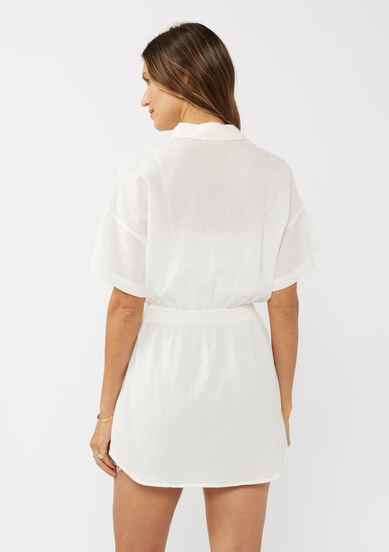 [Color: White] A back facing image of a brunette model wearing a white tunic shirt with a collared neckline, a button front, short sleeves with a dropped shoulder, and a smocked elastic waist. 
