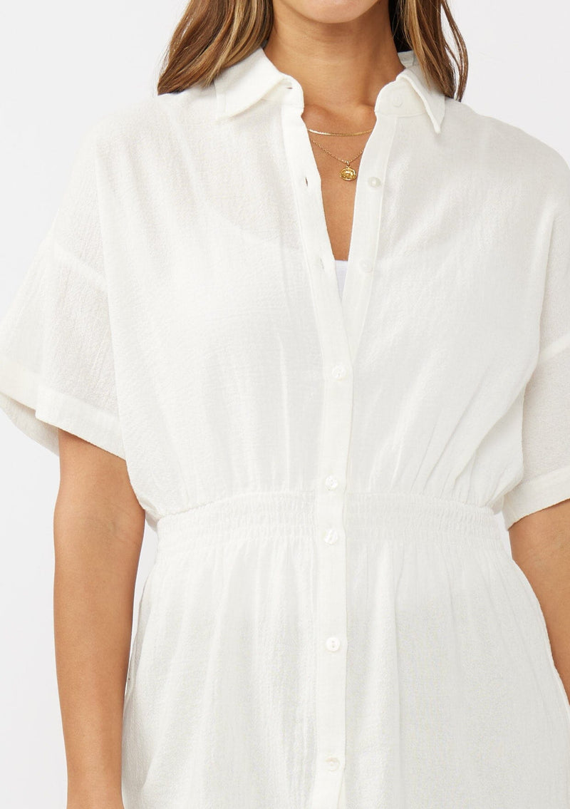 [Color: White] A detail image of a brunette model wearing a white tunic shirt with a collared neckline, a button front, short sleeves with a dropped shoulder, and a smocked elastic waist. 