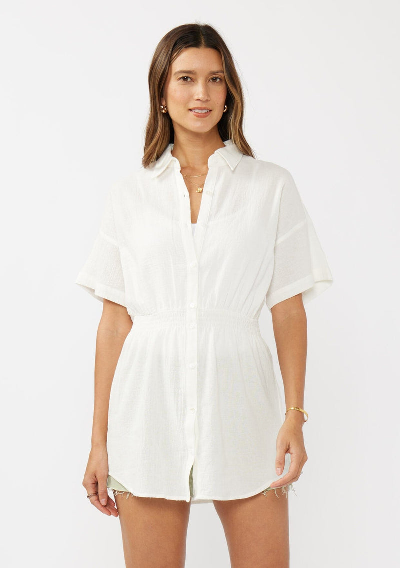 [Color: White] A front facing image of a brunette model wearing a white tunic shirt with a collared neckline, a button front, short sleeves with a dropped shoulder, and a smocked elastic waist. 