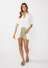 [Color: White] A front facing image of a brunette model wearing a white tunic shirt with a collared neckline, a button front, short sleeves with a dropped shoulder, and a smocked elastic waist. 