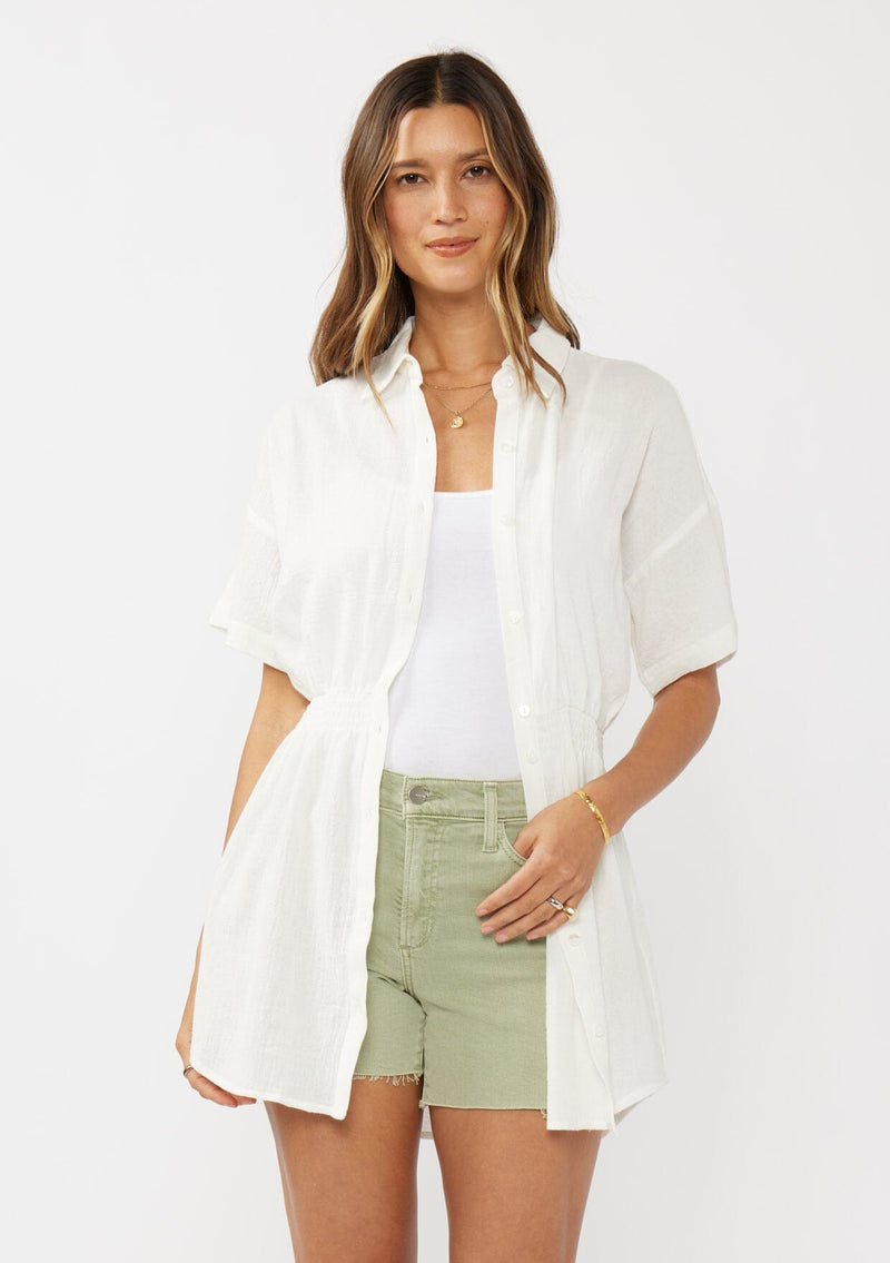 [Color: White] A front facing image of a brunette model wearing a white tunic shirt with a collared neckline, a button front, short sleeves with a dropped shoulder, and a smocked elastic waist. 