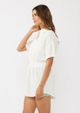[Color: White] A side facing image of a brunette model wearing a white tunic shirt with a collared neckline, a button front, short sleeves with a dropped shoulder, and a smocked elastic waist. 