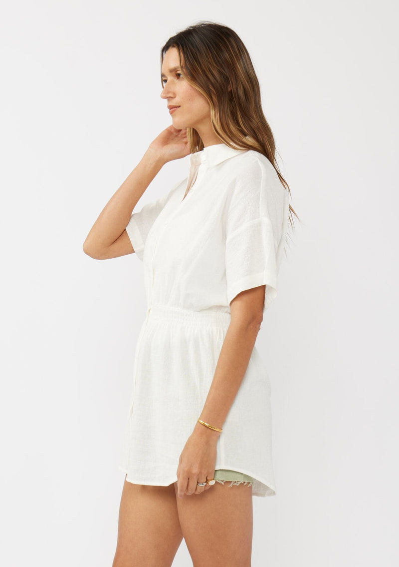 [Color: White] A side facing image of a brunette model wearing a white tunic shirt with a collared neckline, a button front, short sleeves with a dropped shoulder, and a smocked elastic waist. 
