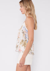 [Color: Off White/Periwinkle] A side facing image of a blonde model wearing a pretty bohemian camisole in an off white and periwinkle blue floral print. With lace trim, adjustable spaghetti straps, a v neckline, and a ruffled hemline. 