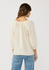[Color: Cream] A back facing image of a brunette model wearing a cream bohemian embroidered blouse with a v-neckline and 3/4 length sleeves with ruffled cuffs. 