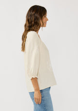 [Color: Cream] A side facing image of a brunette model wearing a cream bohemian embroidered blouse with a v-neckline and 3/4 length sleeves with ruffled cuffs. 