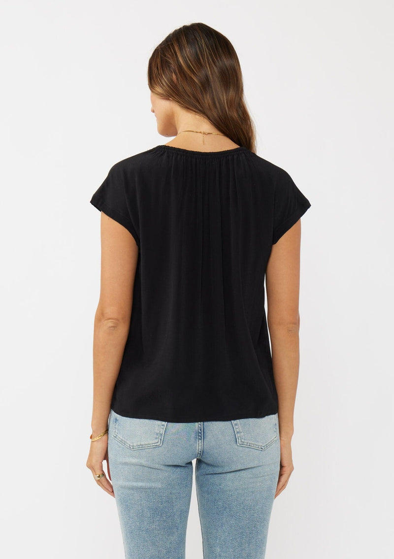[Color: Black] A back facing image of a brunette model wearing a relaxed fit  black top. With self neck ties, ruched split v neckline, and short dolman sleeves. Styled with a cute vintage denim for a laid back look. 