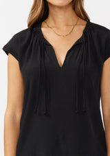 [Color: Black] A detail image of a brunette model wearing a relaxed fit  black top. With self neck ties, ruched split v neckline, and short dolman sleeves. Styled with a cute vintage denim for a laid back look. 