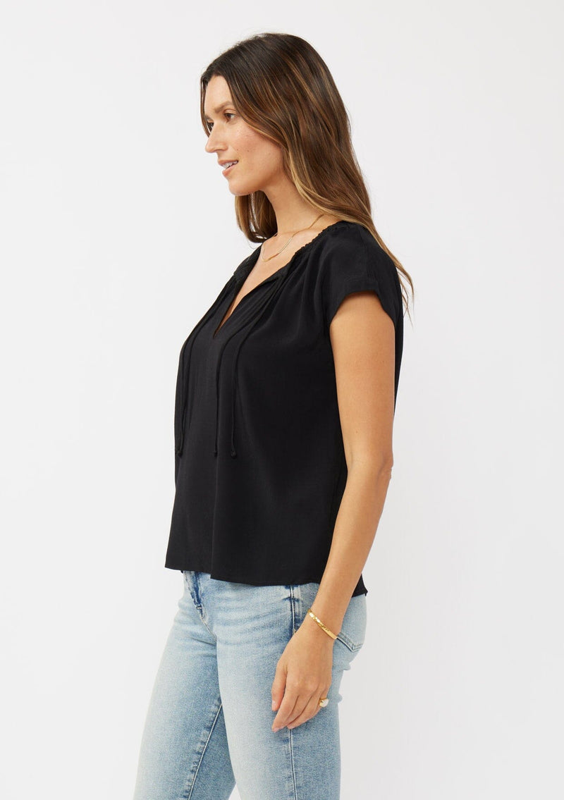 [Color: Black] A side facing image of a brunette model wearing a relaxed fit  black top. With self neck ties, ruched split v neckline, and short dolman sleeves. Styled with a cute vintage denim for a laid back look. 