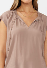 [Color: Mocha] A detail image of a brunette model wearing a relaxed fit  brown mocha top. With self neck ties, ruched split v neckline, and short dolman sleeves. Styled with a cute wide leg denim for a laid back look. 