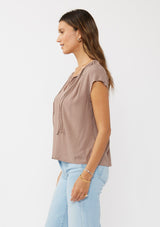 [Color: Mocha] A side facing image of a brunette model wearing a relaxed fit  brown mocha top. With self neck ties, ruched split v neckline, and short dolman sleeves. Styled with a cute wide leg denim for a laid back look. 