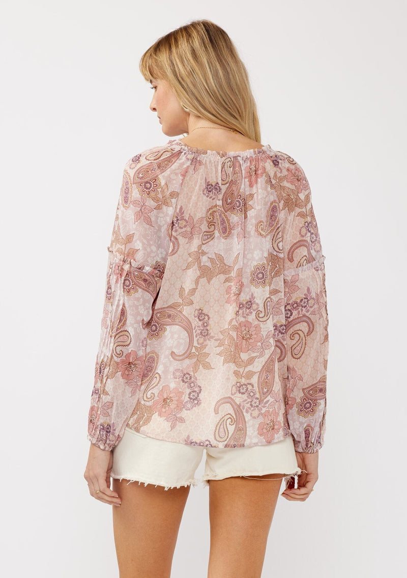 [Color: Blush/Rose] A back facing image of a blonde model wearing a paisley floral print blouse with gold foil accents. Featuring a split v-neckline, long sleeves, a button front, pintuck details, and ruffle trim details. 