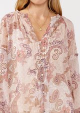 [Color: Blush/Rose] A detail image of a blonde model wearing a paisley floral print blouse with gold foil accents. Featuring a split v-neckline, long sleeves, a button front, pintuck details, and ruffle trim details. 