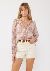 [Color: Blush/Rose] A front facing image of a blonde model wearing a paisley floral print blouse with gold foil accents. Featuring a split v-neckline, long sleeves, a button front, pintuck details, and ruffle trim details. 