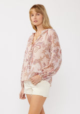 [Color: Blush/Rose] A side facing image of a blonde model wearing a paisley floral print blouse with gold foil accents. Featuring a split v-neckline, long sleeves, a button front, pintuck details, and ruffle trim details. 