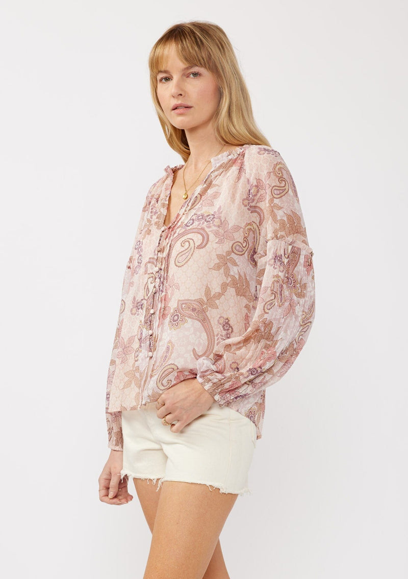 [Color: Blush/Rose] A side facing image of a blonde model wearing a paisley floral print blouse with gold foil accents. Featuring a split v-neckline, long sleeves, a button front, pintuck details, and ruffle trim details. 