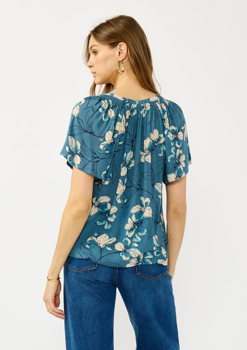 [Color: Teal/Blue] A back facing image of a blonde model wearing a lightweight  teal floral shirt with a split v-neckline and short sleeves. An effortless fall top for casual outings. 