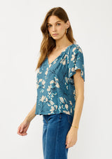 [Color: Teal/Blue] A side facing image of a blonde model wearing a lightweight  teal floral shirt with a split v-neckline and short sleeves. An effortless fall top for casual outings. 