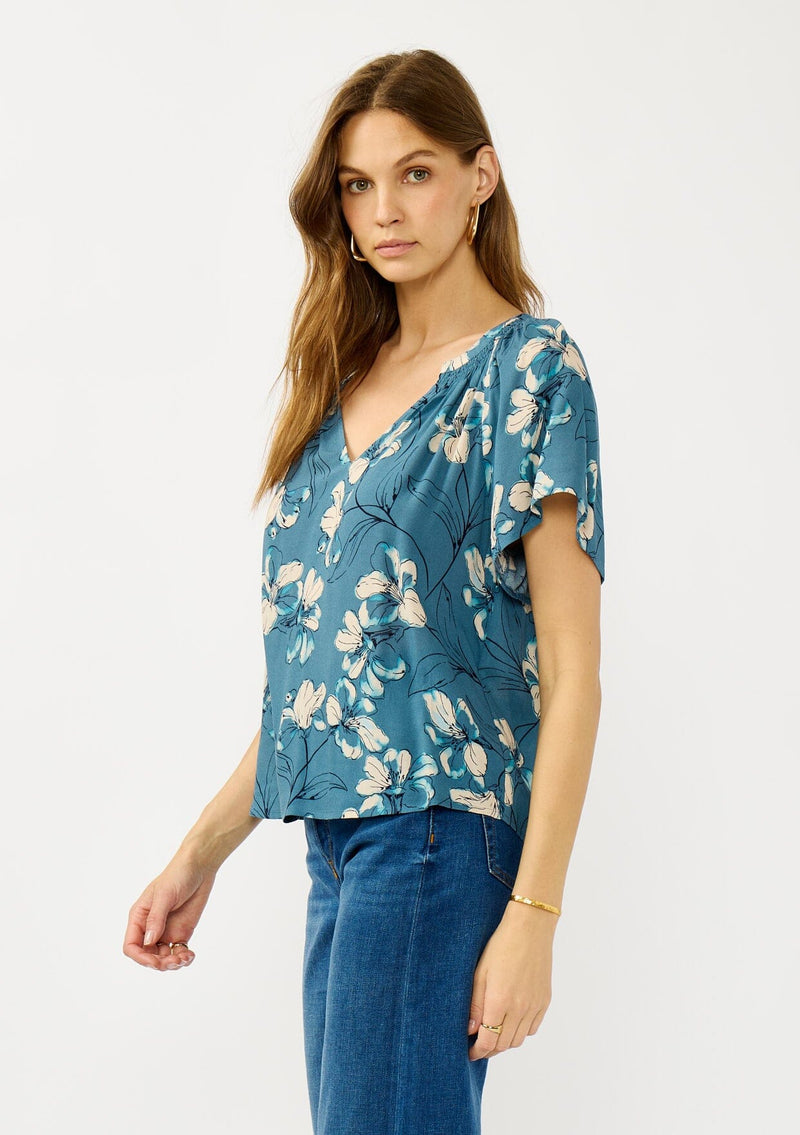 [Color: Teal/Blue] A side facing image of a blonde model wearing a lightweight  teal floral shirt with a split v-neckline and short sleeves. An effortless fall top for casual outings. 