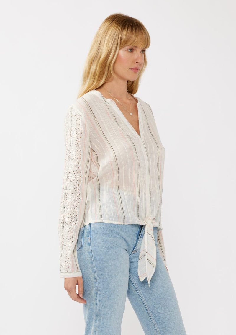 [Color: White/Coral] A side facing image of a blonde model wearing an off white yarn dye stripe top with embroidered eyelet details. This sheer  cotton top features a split v-neckline, long sleeves, button cuff closures, and a tie front hem detail. Perfect for vacation and resort styling. 