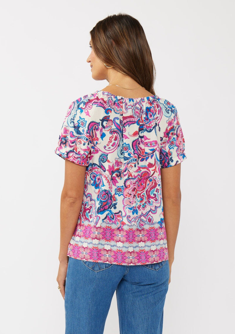 [Color: Natural/Dusty Pink] A back facing image of a brunette model wearing a vibrant floral print boho top with an elastic neckline and short puff sleeves.