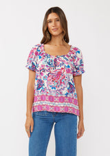 [Color: Natural/Dusty Pink] A front facing image of a brunette model wearing a vibrant floral print boho top with an elastic neckline and short puff sleeves.