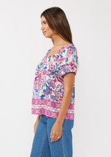 [Color: Natural/Dusty Pink] A side  facing image of a brunette model wearing a vibrant floral print boho top with an elastic neckline and short puff sleeves.