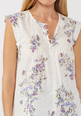 [Color: Off White/Lilac] A detail image of a blonde model wearing a white top adorned with a purple floral print and embroidered eyelet accents. This spring top features a button-front neck closure and delicate pintuck details.