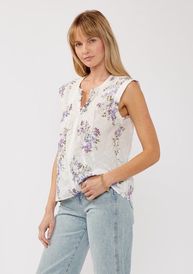 [Color: Off White/Lilac] A side-facing image of a blonde model wearing a white top adorned with a purple floral print and embroidered eyelet accents. This spring top features a button-front neck closure and delicate pintuck details.